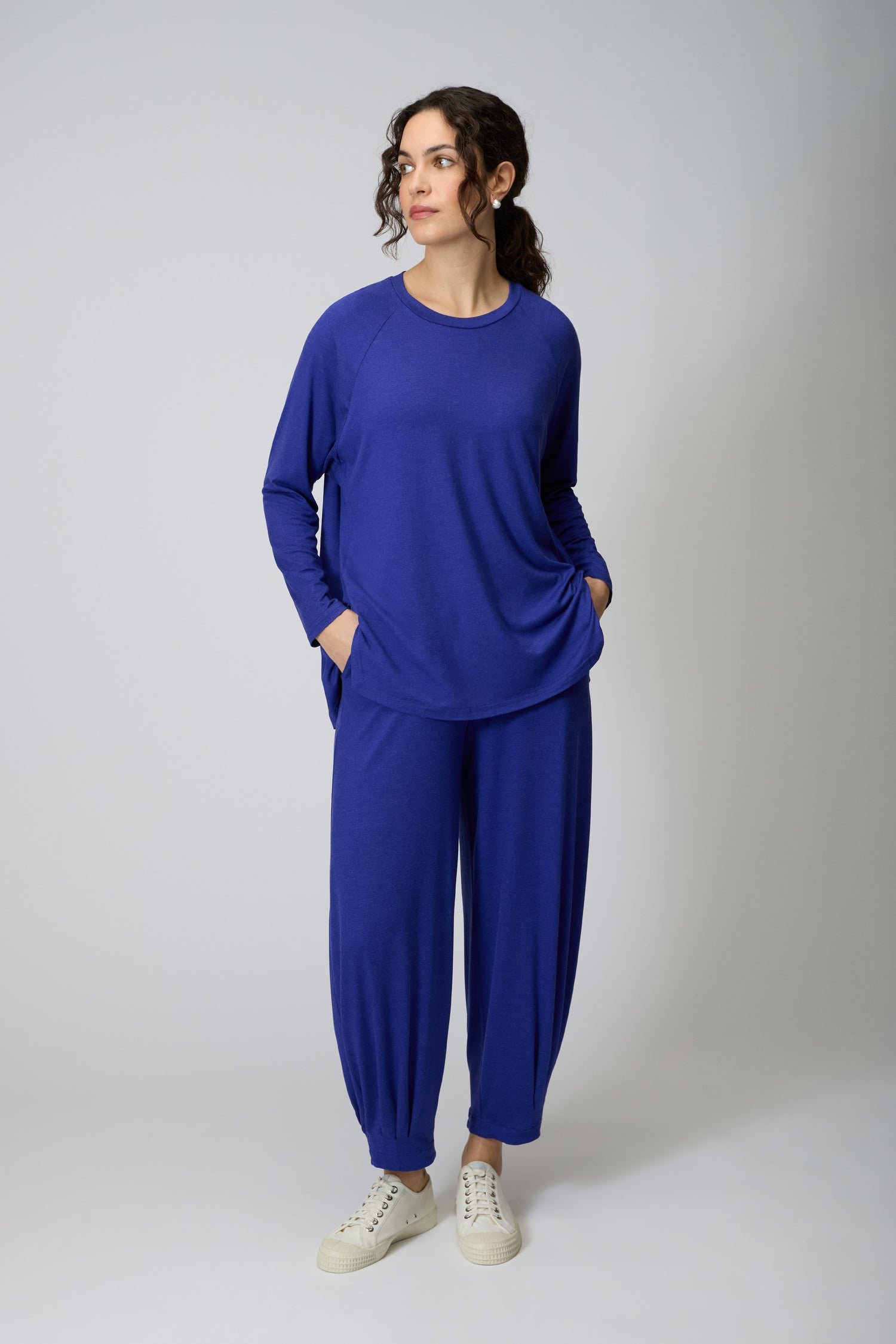 A woman stands against a plain background wearing the Soft Viscose Jersey Boxy Top in blue with matching pants and white sneakers. Her hands are in her pockets as she looks to the side.