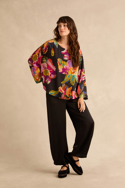A person wears a Pixelated Floral Relaxed Top with a black background and a large, colorful, pixelated floral print, paired with wide-leg black trousers and black Mary Jane flats.