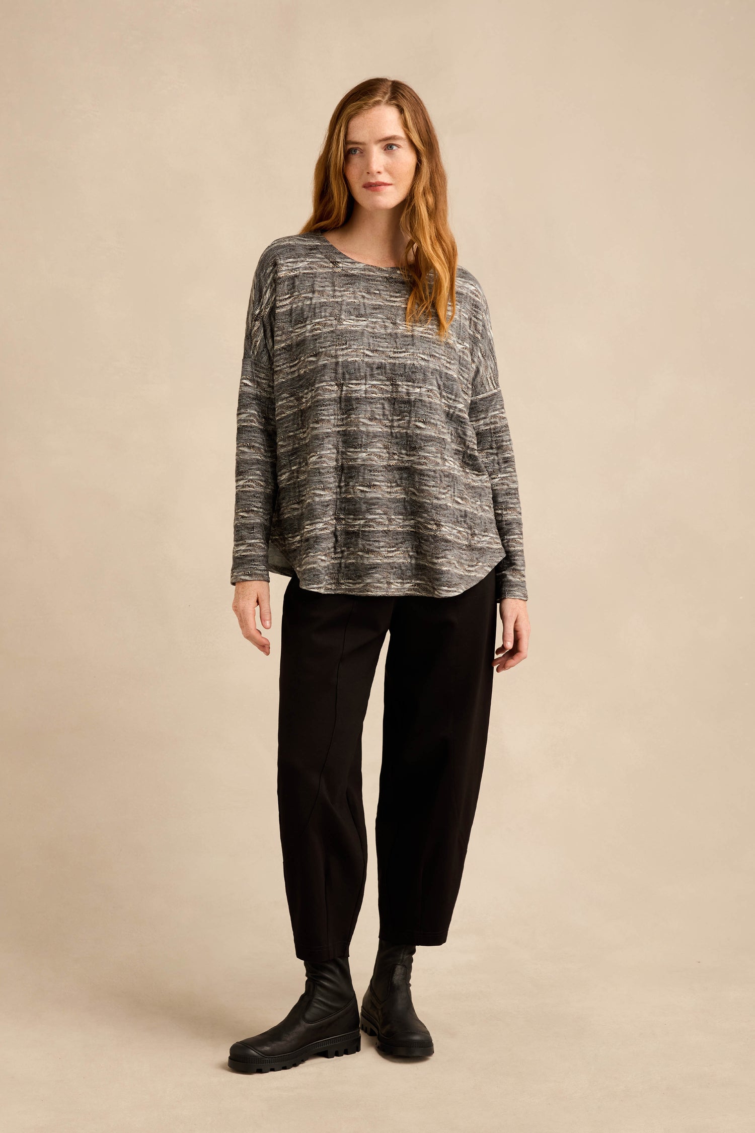 A woman stands facing forward against a plain beige background, wearing the Abstract Ripple Jersey Boxy Top in gray patterns, paired with black pants and black boots.