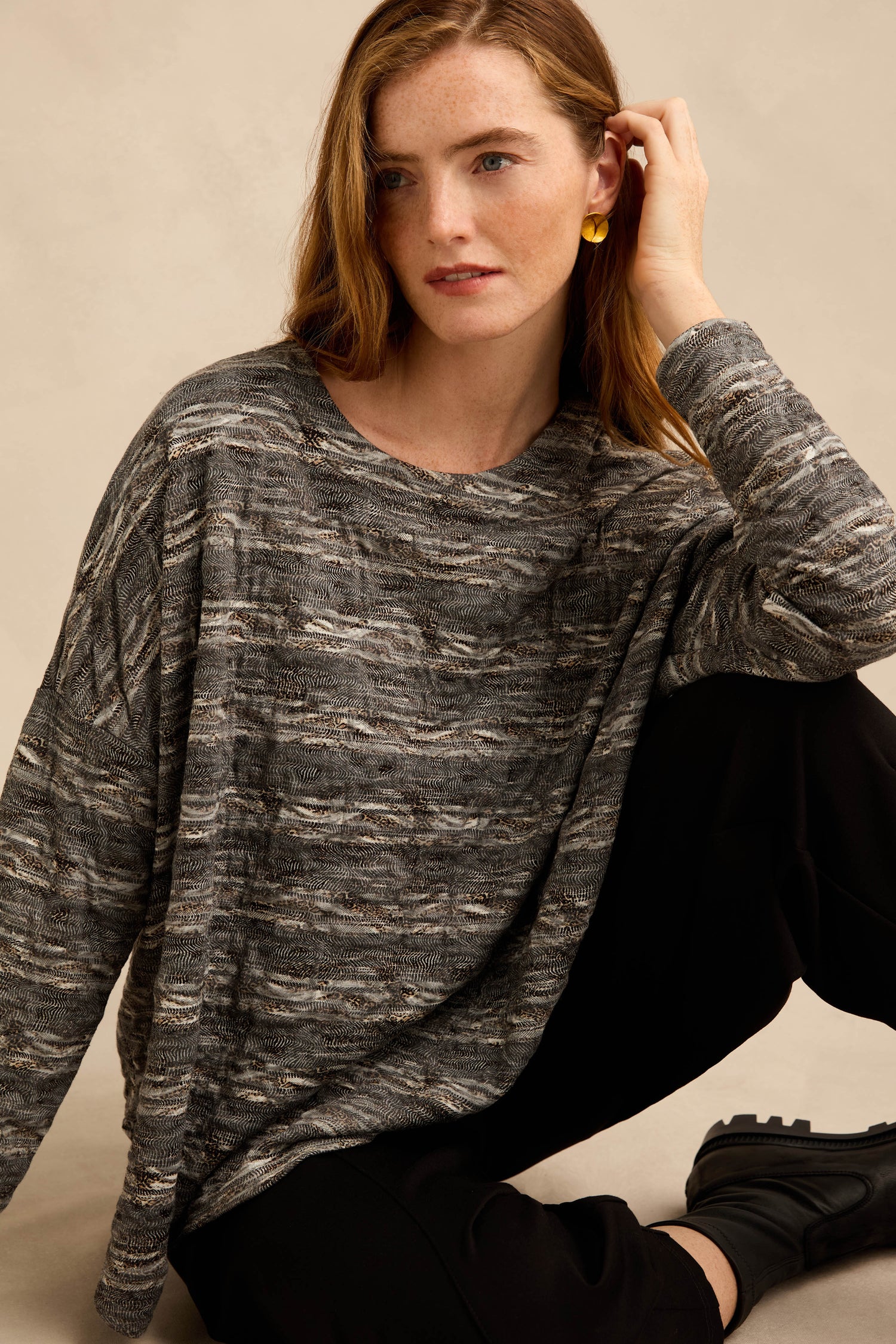 A woman with long hair wears the Abstract Ripple Jersey Boxy Top, featuring a gray patterned design, paired with black pants, black boots, and gold earrings. She sits with one knee up against a neutral background.