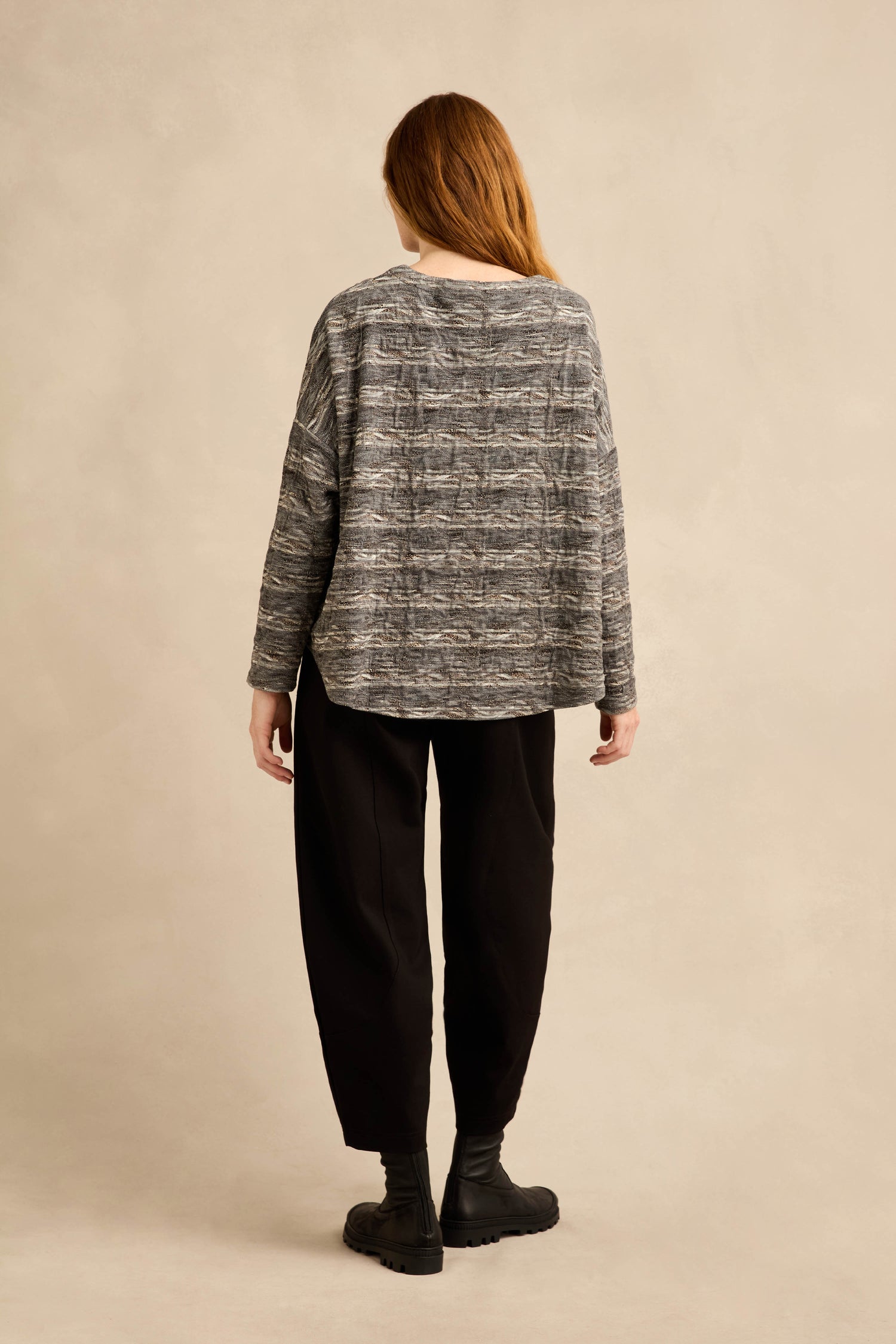 A person with long hair stands facing away, wearing the Abstract Ripple Jersey Boxy Top—loose and gray with a textured finish—paired with black pants and black boots against a plain beige background.