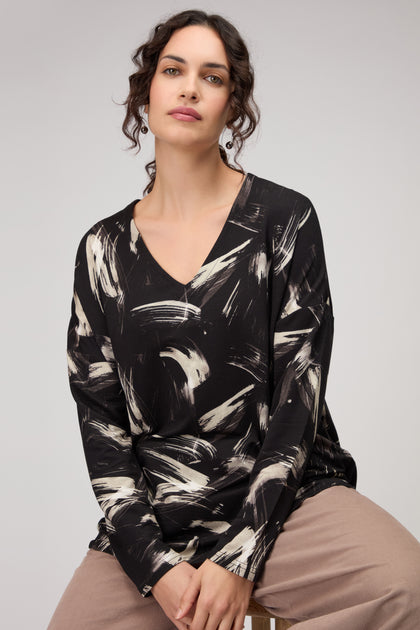 A woman with dark wavy hair wears the Painted Brushstrokes Jersey Top in black and white, paired with beige pants, as she sits on a stool against a plain background.