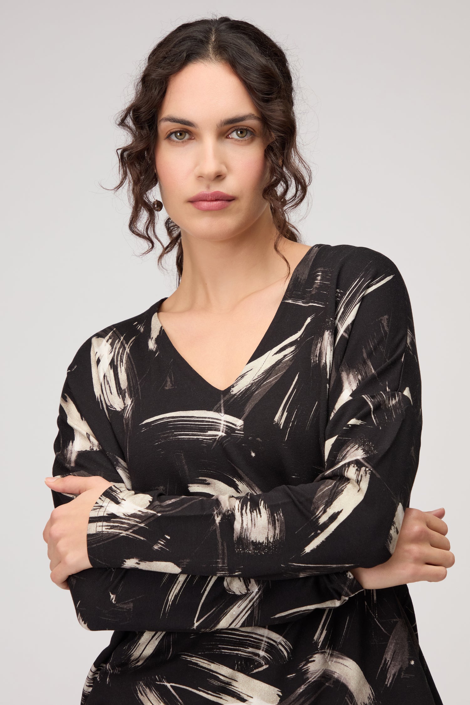 Woman with wavy dark hair wears the Painted Brushstrokes Jersey Top, a black V-neck women’s jersey featuring beige painted brushstrokes, standing with arms crossed against a plain background.
