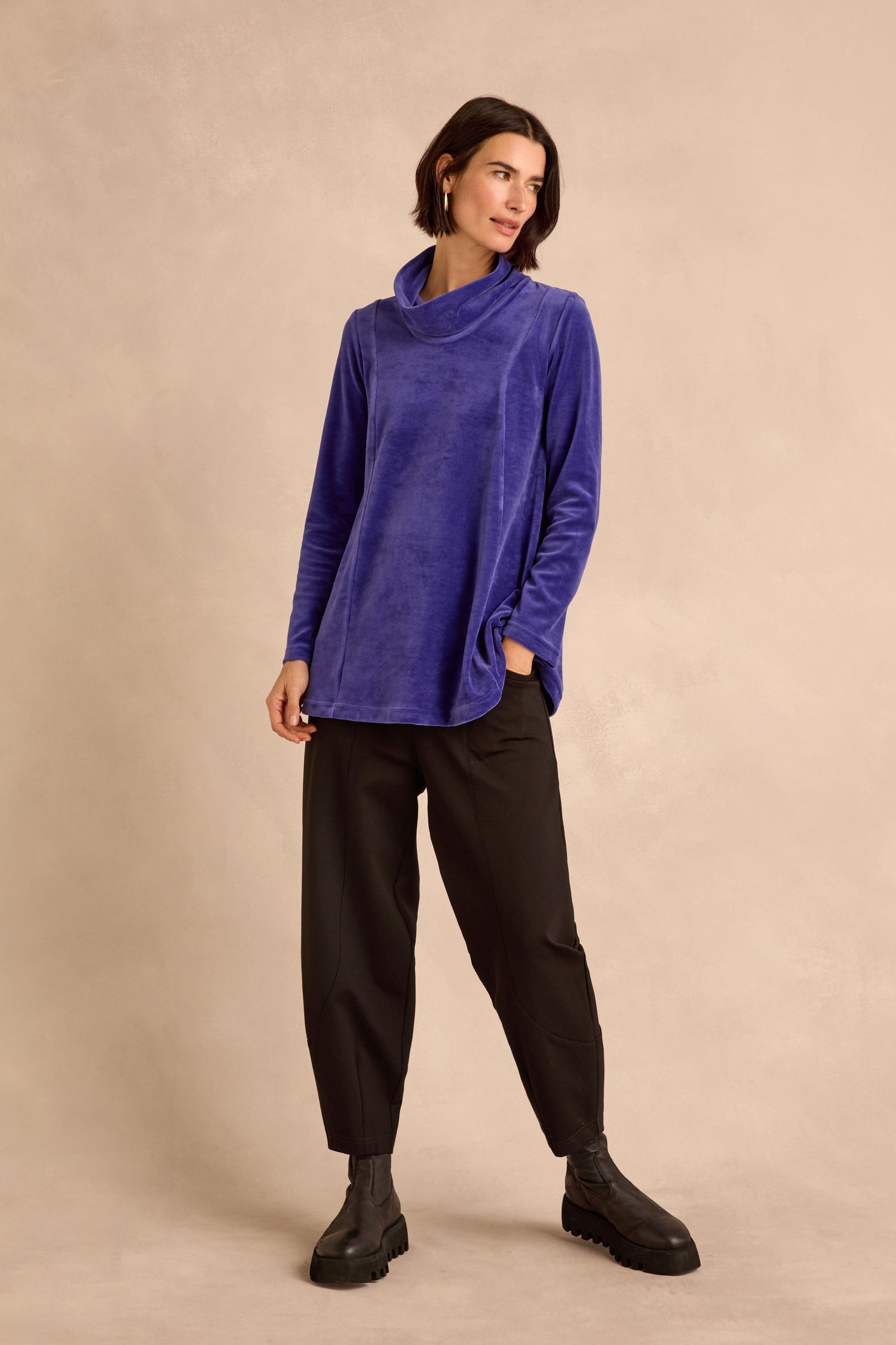 A woman stands in front of a beige background wearing the Velvet Jersey Cowl Neck Top—an effortlessly stylish, long-sleeve blue velvet jersey layer perfect for winter—paired with loose black pants and black ankle boots, one hand casually tucked in her pocket.