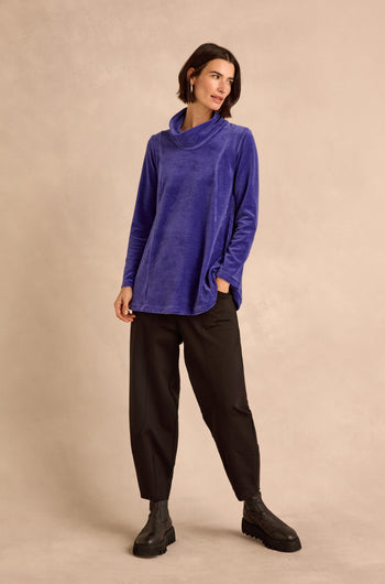 A woman stands in front of a beige background wearing the Velvet Jersey Cowl Neck Top—an effortlessly stylish, long-sleeve blue velvet jersey layer perfect for winter—paired with loose black pants and black ankle boots, one hand casually tucked in her pocket.
