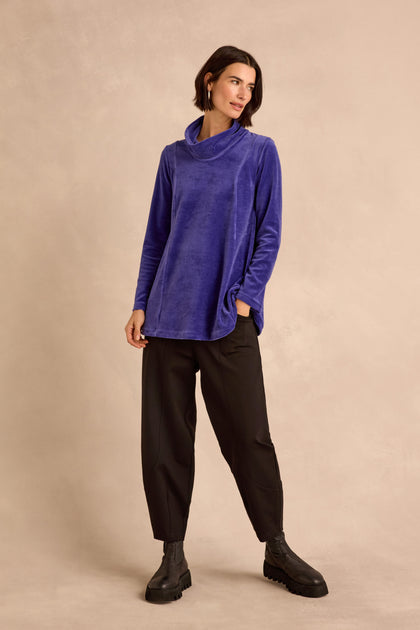 A woman stands in front of a beige background wearing the Velvet Jersey Cowl Neck Top—an effortlessly stylish, long-sleeve blue velvet jersey layer perfect for winter—paired with loose black pants and black ankle boots, one hand casually tucked in her pocket.