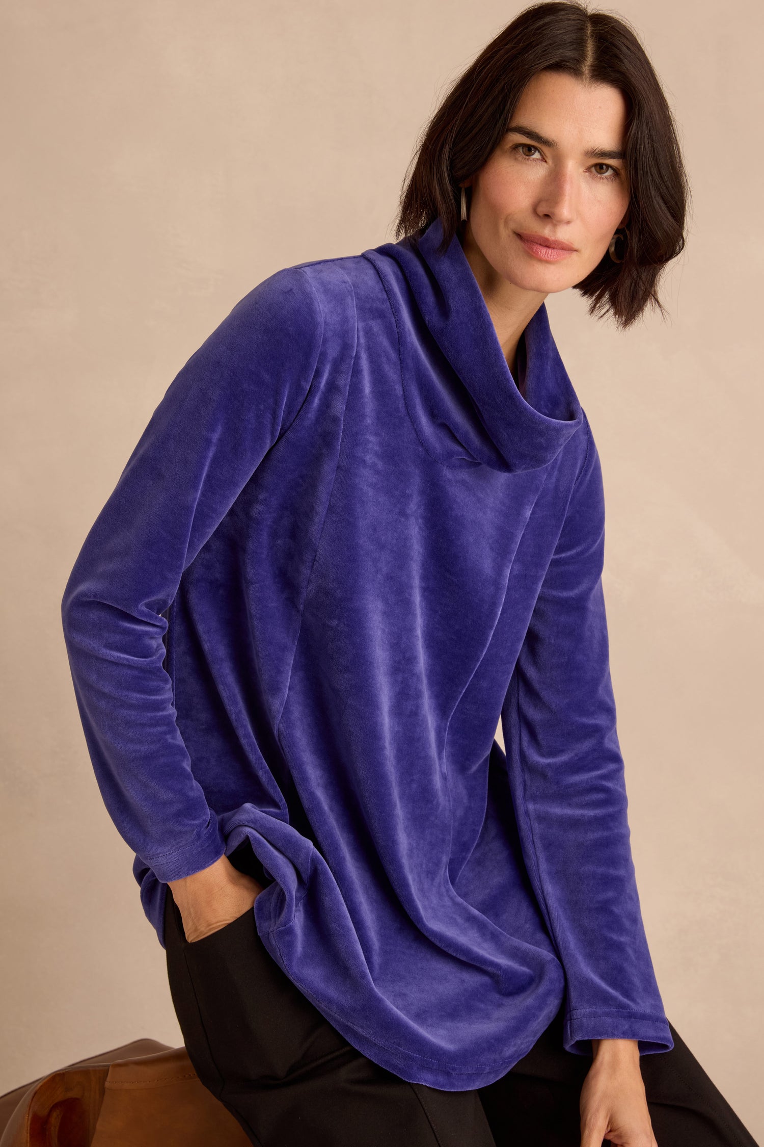A woman with short dark hair wears the Velvet Jersey Cowl Neck Top—a loose-fitting, long-sleeve blue style perfect for winter layering—paired with black pants, seated with one hand in her pocket and looking at the camera.