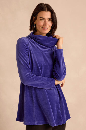 A woman smiles at the camera while standing against a neutral background, wearing the Velvet Jersey Cowl Neck Top in long-sleeve blue velvet—a perfect winter layer.