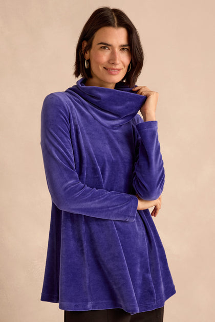 A woman smiles at the camera while standing against a neutral background, wearing the Velvet Jersey Cowl Neck Top in long-sleeve blue velvet—a perfect winter layer.