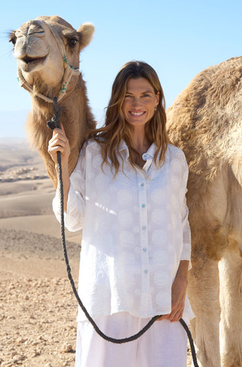 A woman wears the Jacquard Circles Shirt, an oversized white shirt, as she smiles in a desert holding a camel by its lead. Textured circles accent her outfit, and part of the camel is seen beside her.