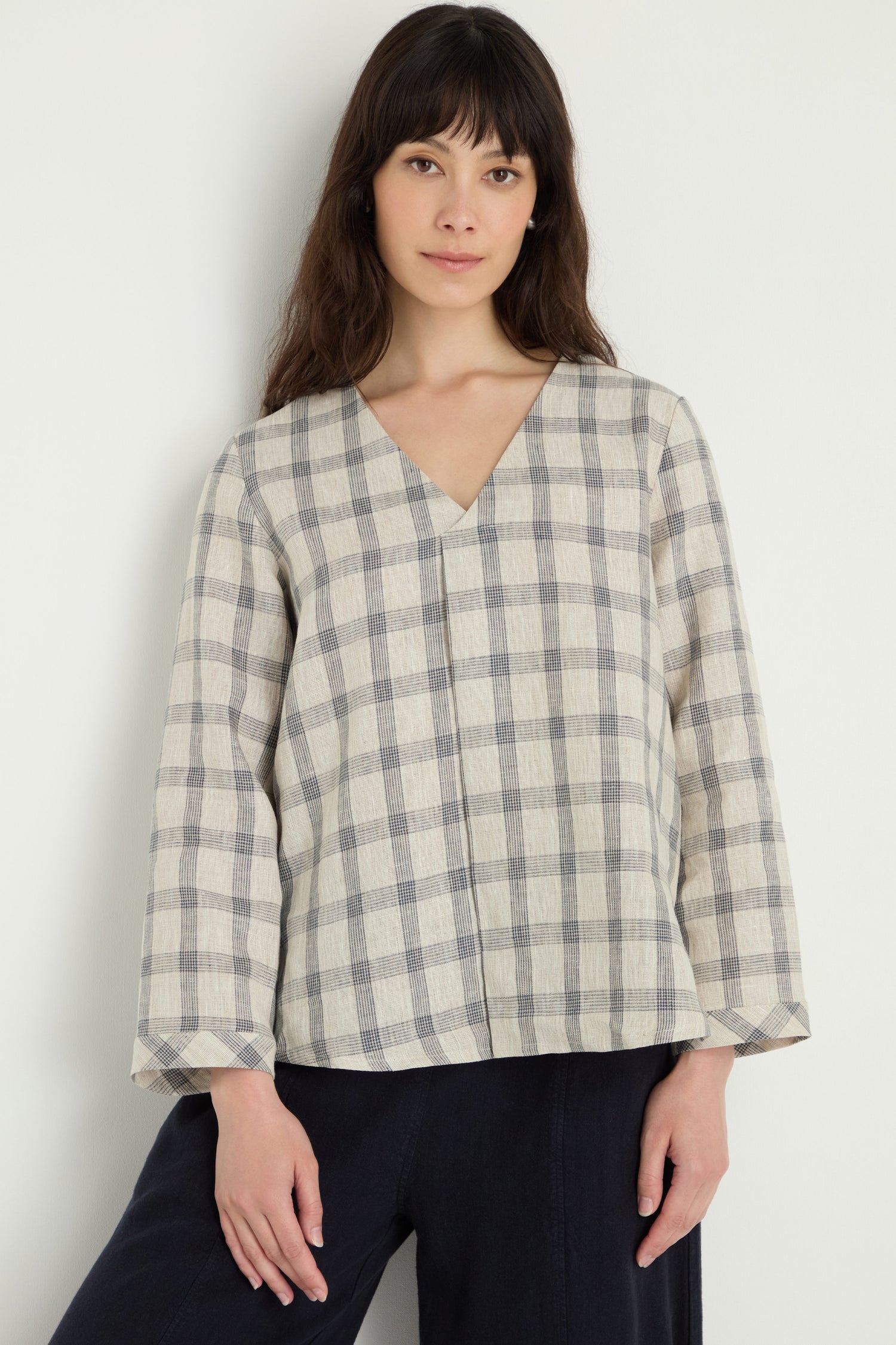 A woman stands against a white wall wearing the Grid Check Linen Top—a relaxed fit, long-sleeve V-neck blouse in beige and gray grid check—paired with loose black pants.