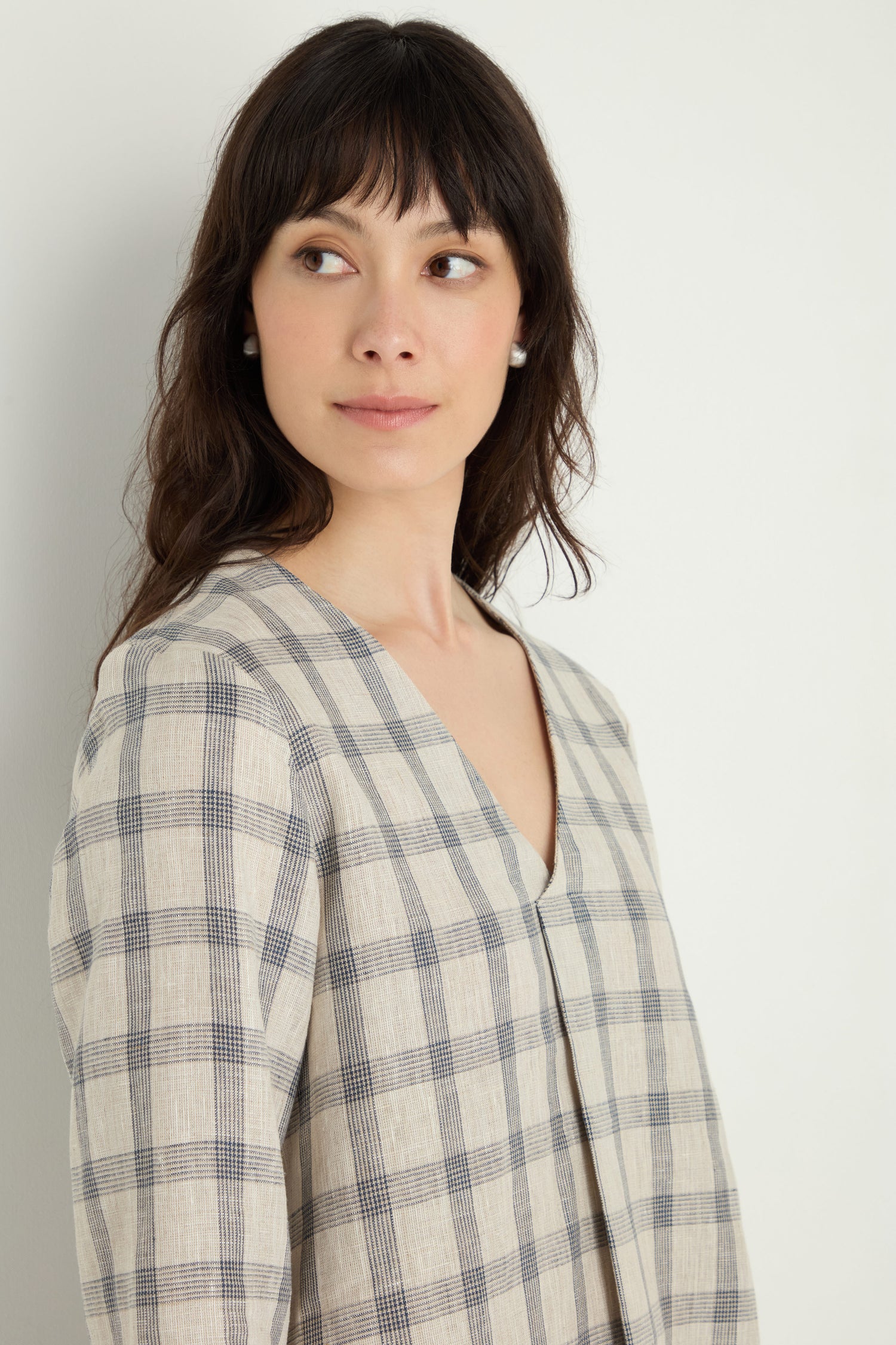 A woman with long dark hair models the Grid Check Linen Top in a relaxed fit, standing against a plain white background and gazing slightly to her right.