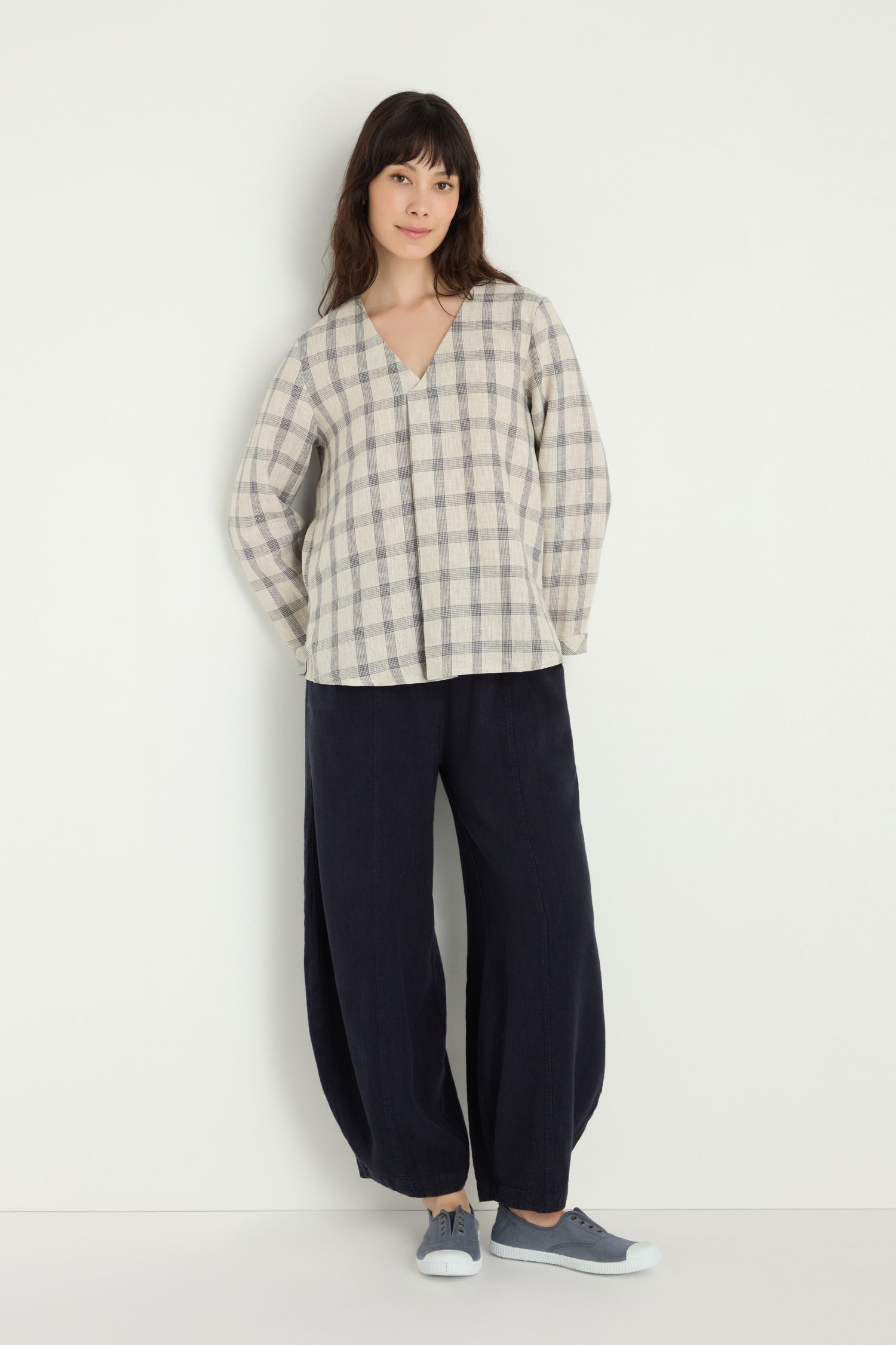 A woman stands against a white wall wearing the Grid Check Linen Top, loose dark pants, and gray sneakers.