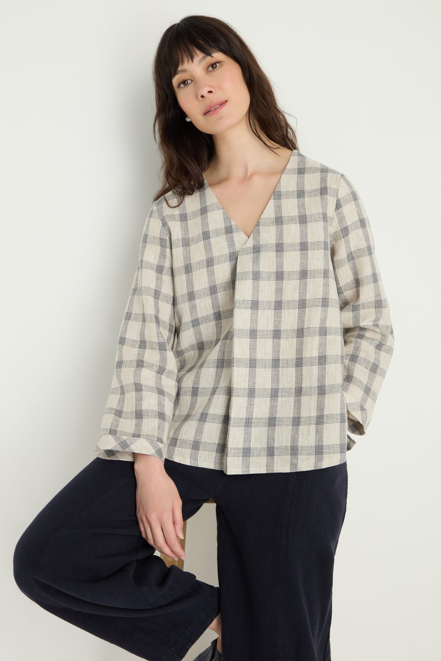 A woman wears the Grid Check Linen Top, a relaxed-fit, long-sleeve beige and gray blouse, paired with dark pants against a plain light background.