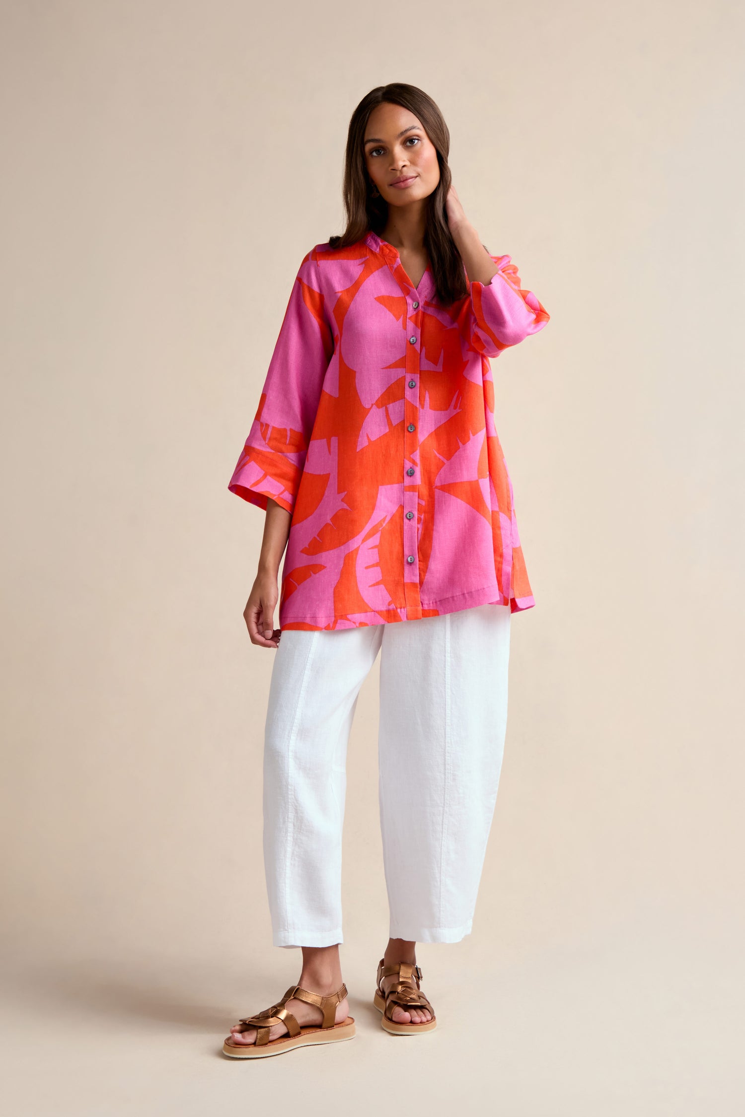 A woman stands against a plain background wearing the Palm Mirage Linen Shirt, featuring a bright pink and orange tropical print with a relaxed fit, paired with white loose pants and brown sandals.