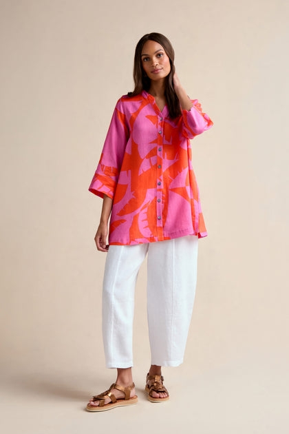 A woman stands against a plain background wearing the Palm Mirage Linen Shirt, featuring a bright pink and orange tropical print with a relaxed fit, paired with white loose pants and brown sandals.