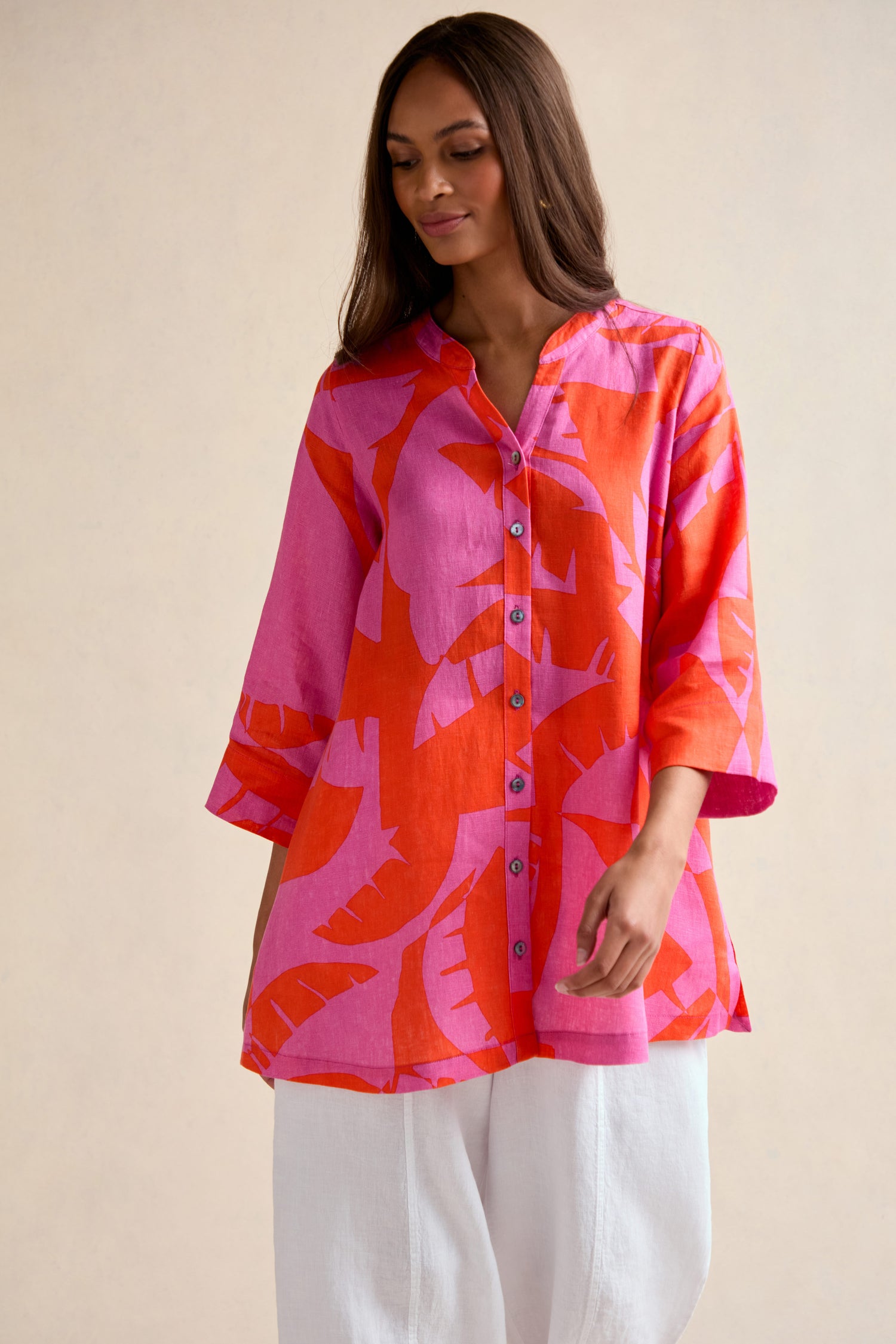 A woman wears the Palm Mirage Linen Shirt, featuring a relaxed fit and bold pink and orange tropical print, paired with white pants against a neutral background.