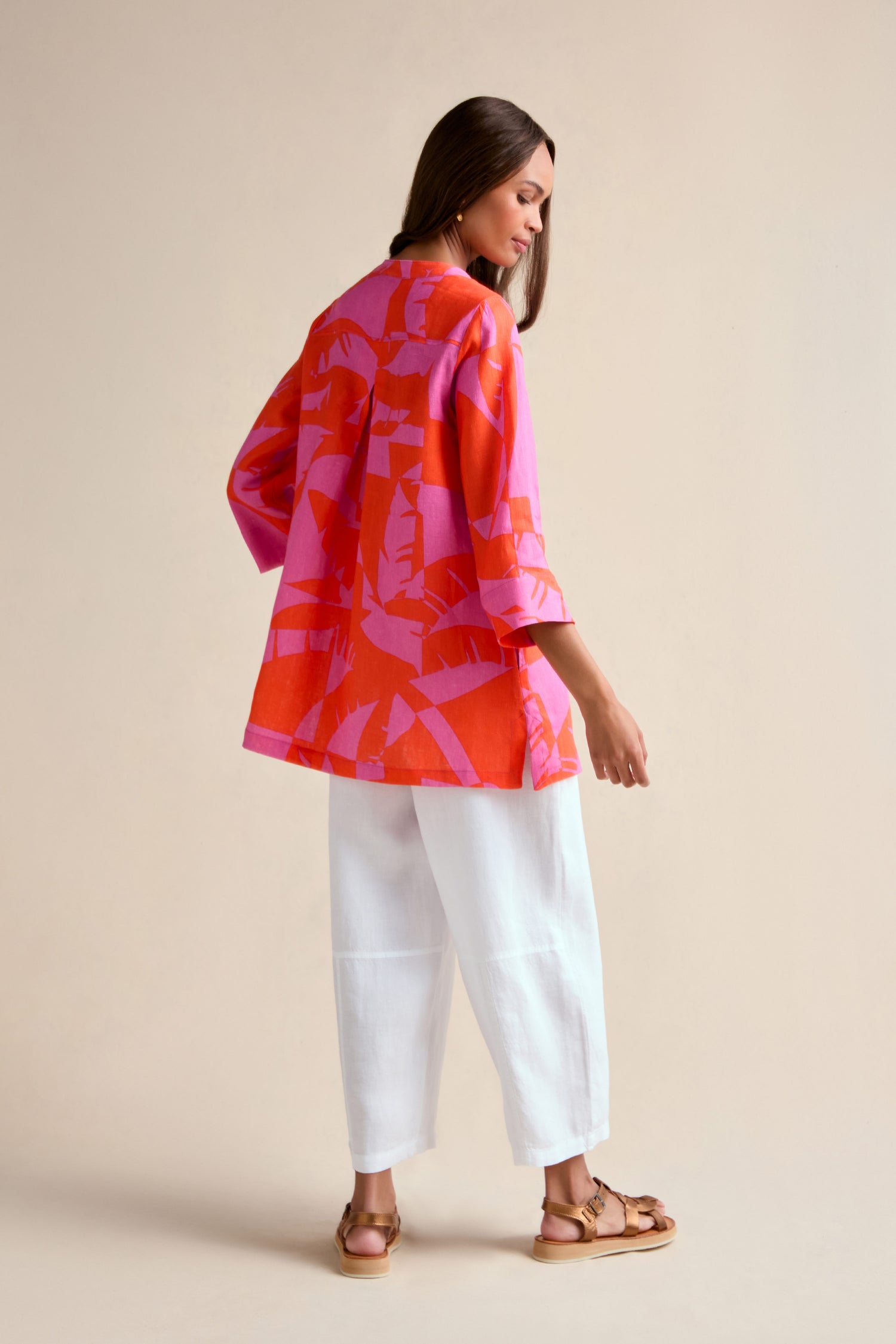 A woman stands against a plain light background, looking over her shoulder in the Palm Mirage Linen Shirt with a tropical pink and red print, paired with white cropped pants and sandals.