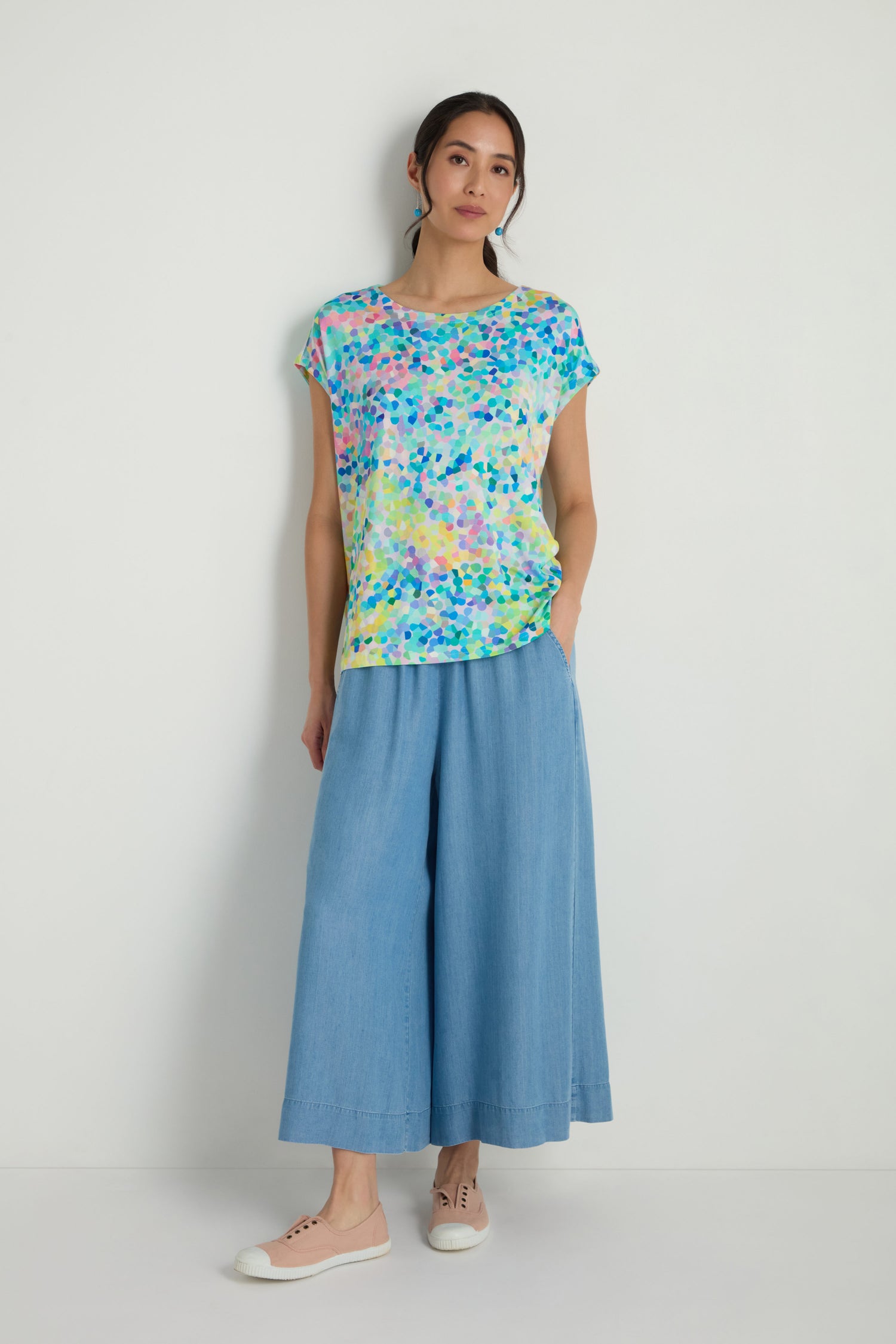 A woman stands against a white wall wearing the Summer Confetti Jersey Top, featuring a boxy silhouette, paired with wide-leg light blue pants and beige sneakers.