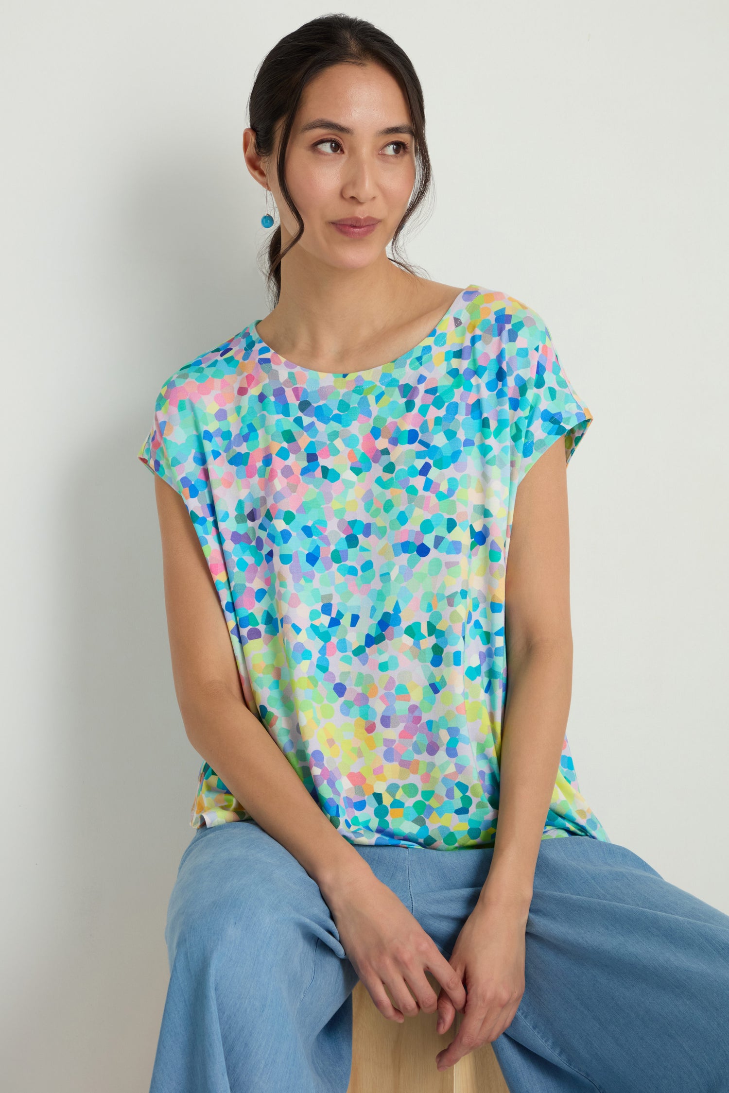 A woman with her hair tied back sits on a stool, wearing the Summer Confetti Jersey Top in an oversized fit and light blue pants, looking slightly to the side against a plain background.