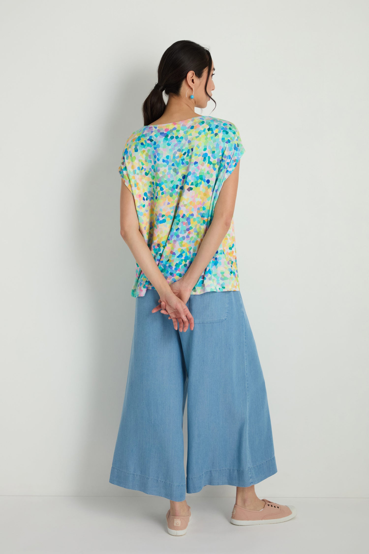 A woman stands against a white wall, facing away with her hands clasped behind her back, wearing an oversized Summer Confetti Jersey Top, wide-leg light blue jeans, and beige shoes.