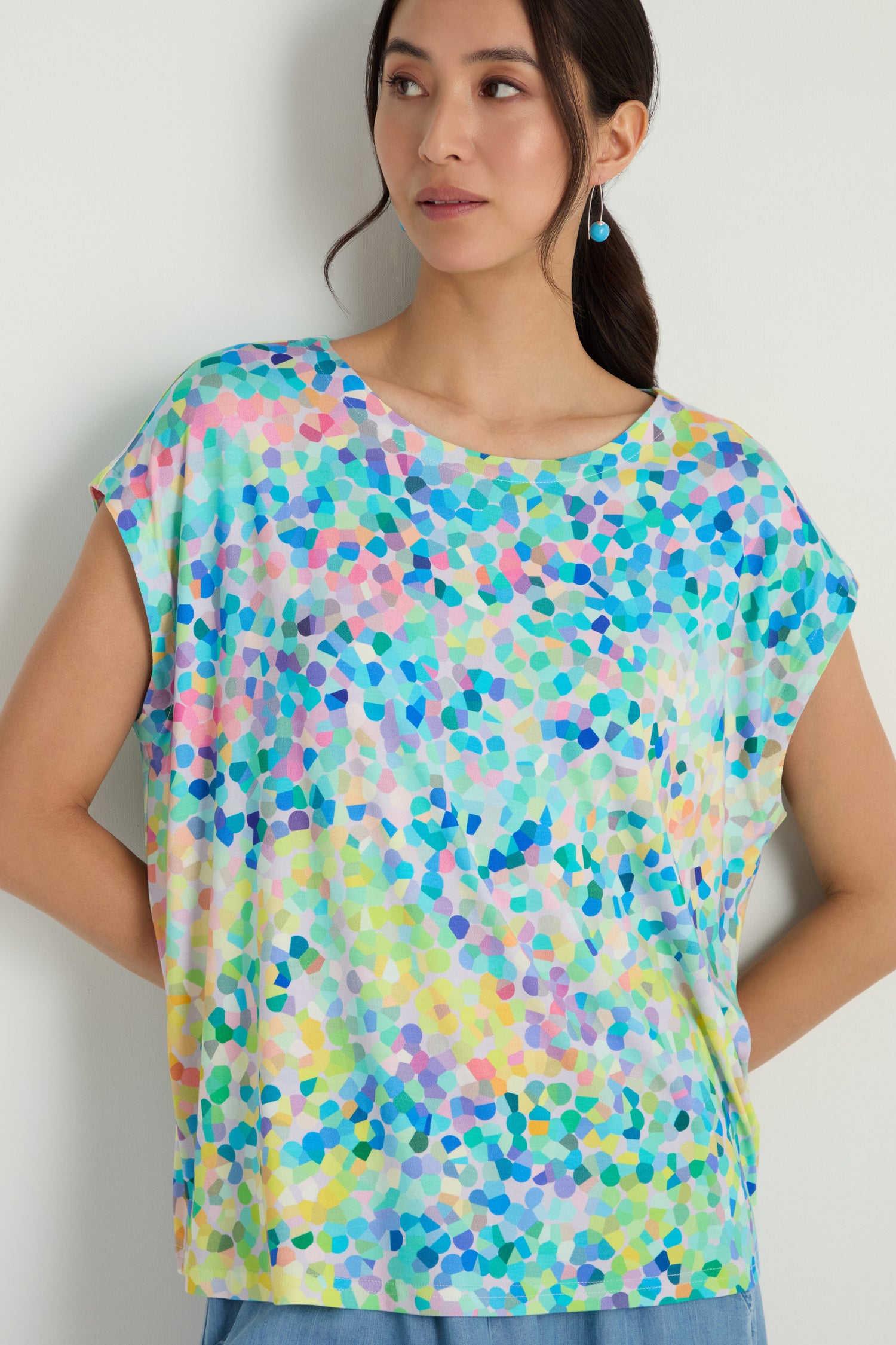 A woman models the Summer Confetti Jersey Top, featuring a colorful abstract mosaic pattern and boxy silhouette, posed against a plain light-colored wall.