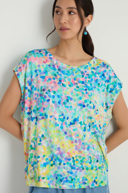 A woman models the Summer Confetti Jersey Top, featuring a colorful abstract mosaic pattern and boxy silhouette, posed against a plain light-colored wall.