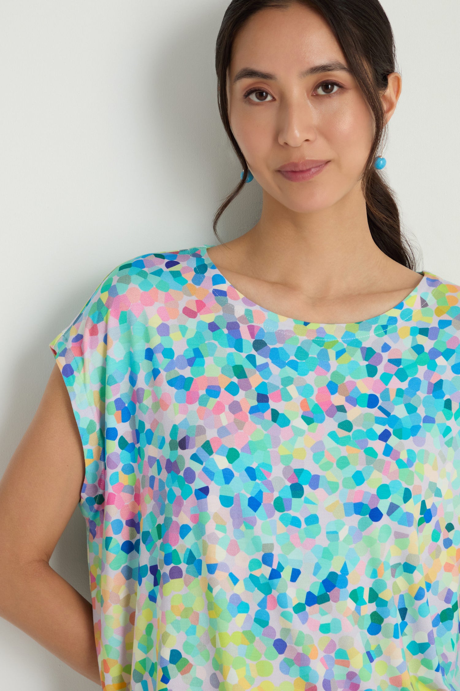 A woman with long dark hair in a ponytail, wearing blue earrings and the oversized Summer Confetti Jersey Top, stands against a plain light background.