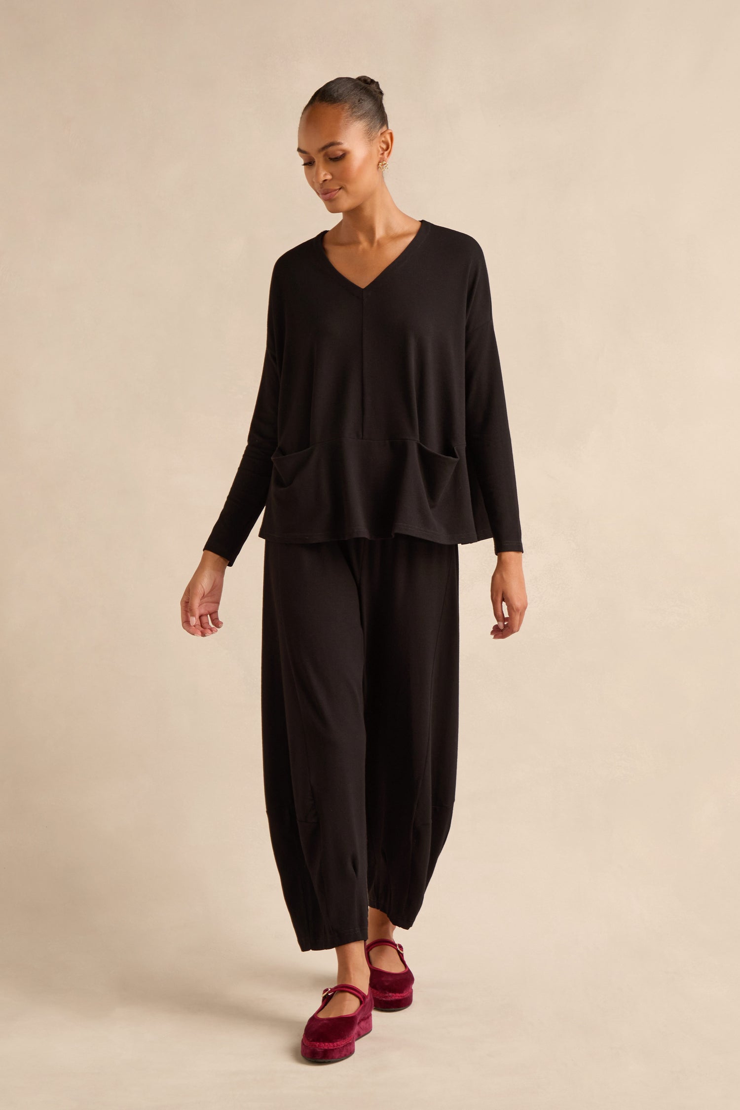 A person stands against a neutral beige background, wearing the Fluid Crepe Relaxed Top—an easy black V-neck crepe piece with two front pockets and a boxy silhouette—paired with matching wide-leg black pants and red shoes.