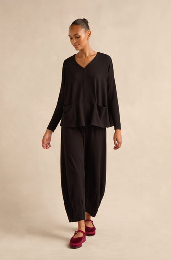 A person stands against a neutral beige background, wearing the Fluid Crepe Relaxed Top—an easy black V-neck crepe piece with two front pockets and a boxy silhouette—paired with matching wide-leg black pants and red shoes.