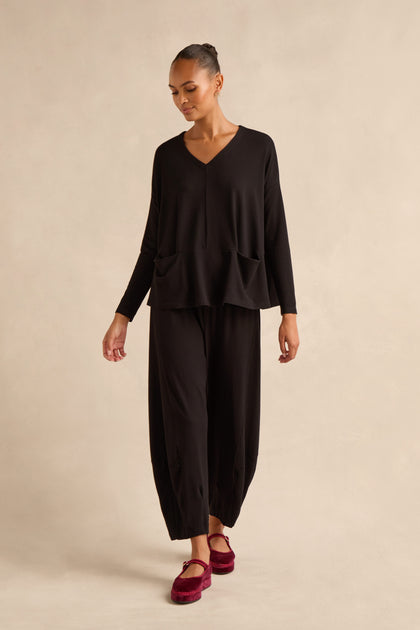 A person stands against a neutral beige background, wearing the Fluid Crepe Relaxed Top—an easy black V-neck crepe piece with two front pockets and a boxy silhouette—paired with matching wide-leg black pants and red shoes.