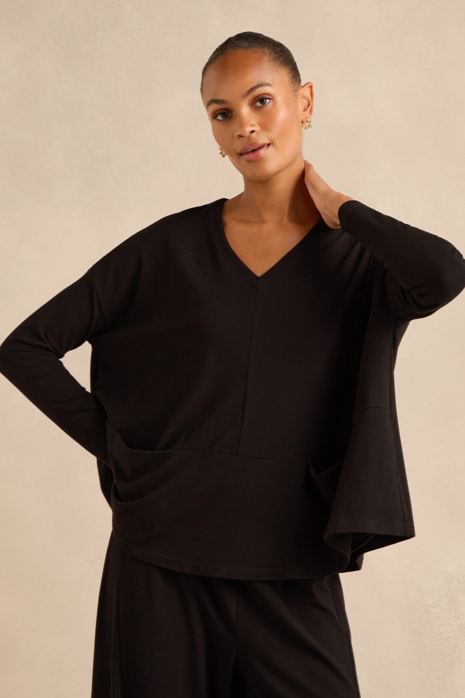 A woman wearing the Fluid Crepe Relaxed Top in black, styled with matching black pants and standing against a plain beige background.