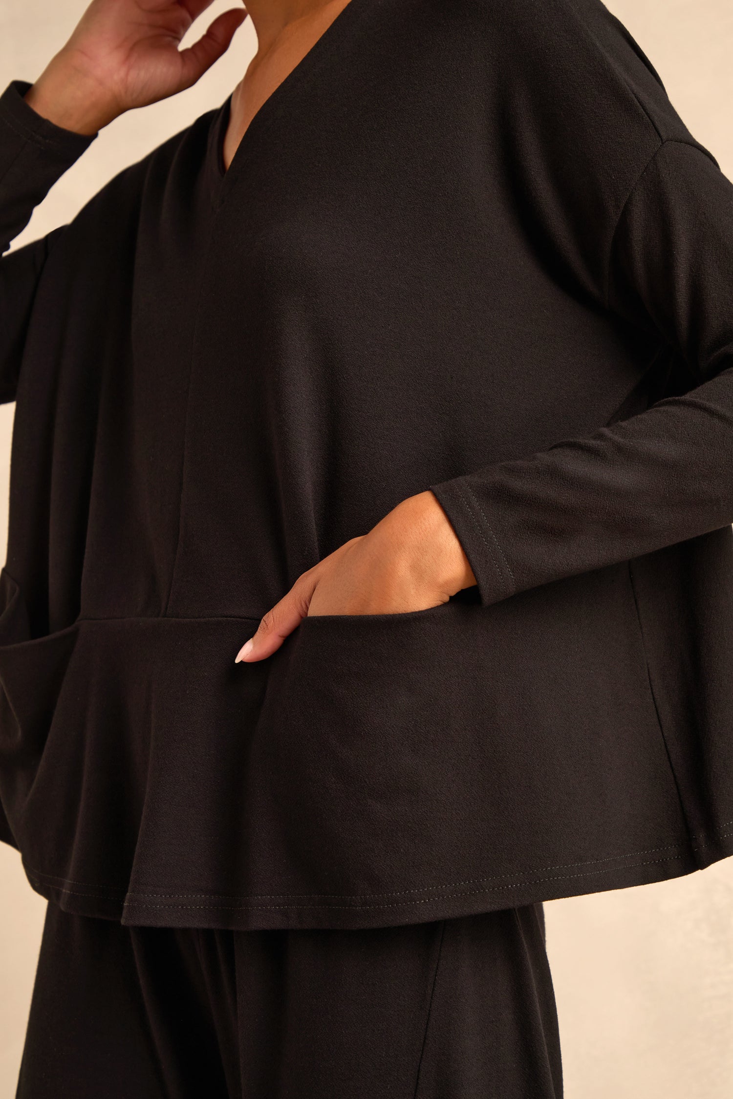 A person wearing the Fluid Crepe Relaxed Top in black—a loose-fitting, boxy silhouette—paired with black pants, one hand resting in a side pocket and the other touching their neck.