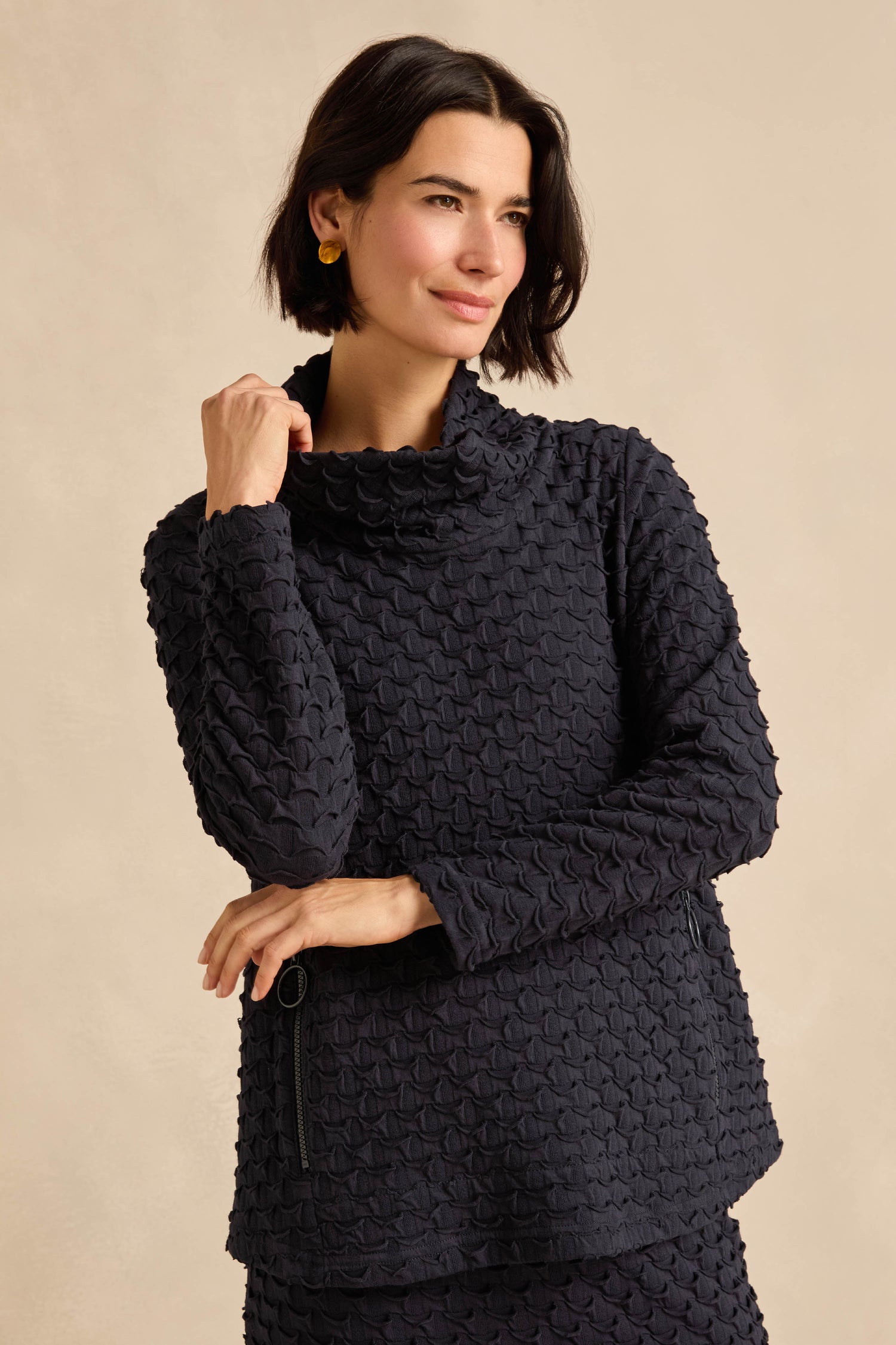 Person with short dark hair wearing a textured stretch jersey, long-sleeve dark top—such as the Origami Pocket Jersey Cowl Neck Top, made in England—standing against a neutral background with one hand touching their chin.