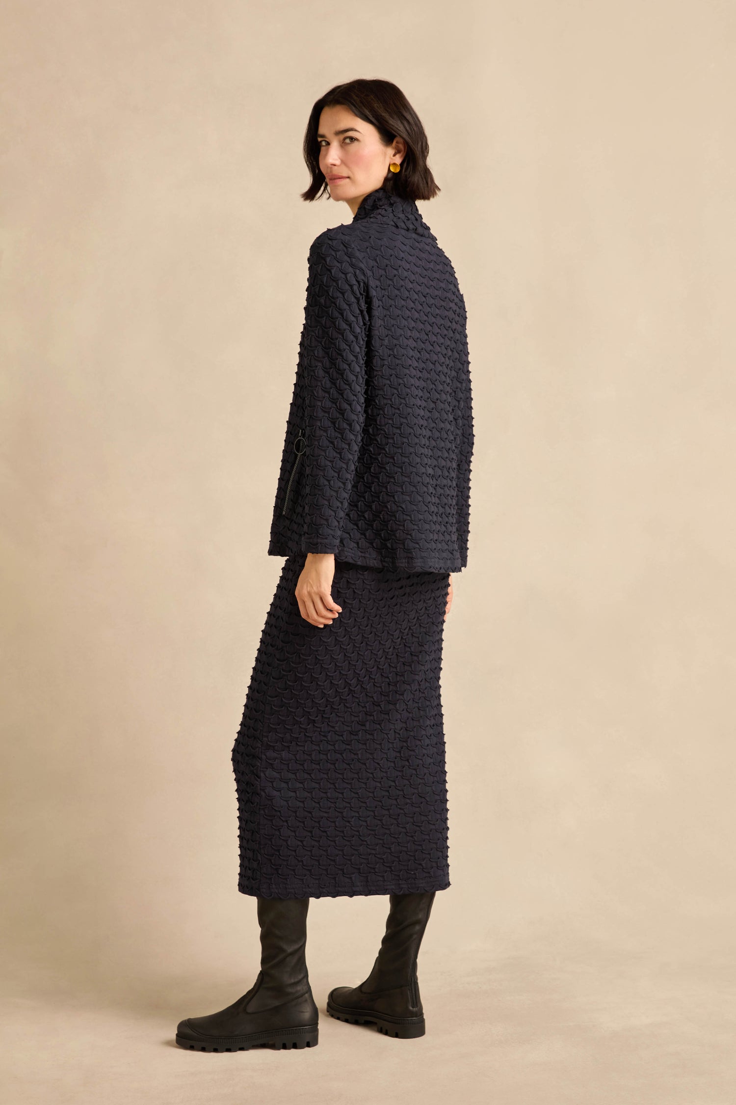 Woman standing sideways, wearing the Origami Pocket Jersey Cowl Neck Top with a matching skirt and black boots, against a plain beige background.