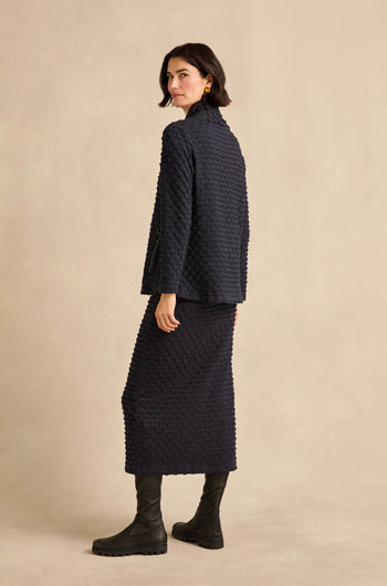 Woman standing sideways, wearing the Origami Pocket Jersey Cowl Neck Top with a matching skirt and black boots, against a plain beige background.