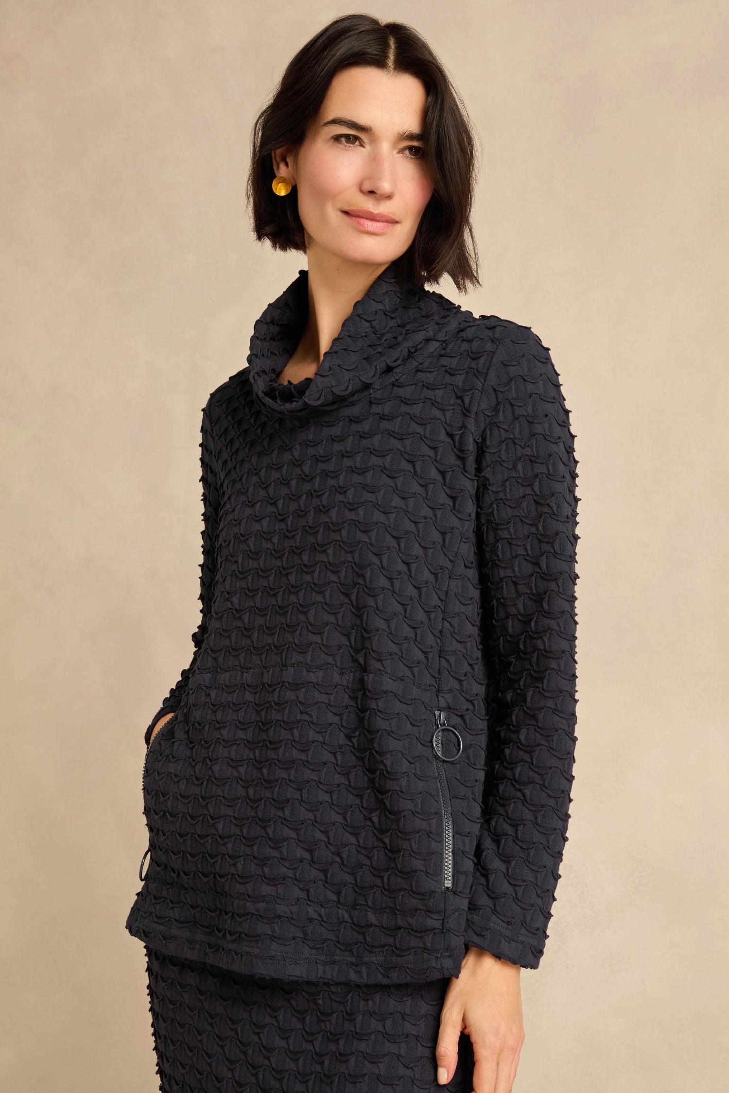 Woman wearing the Origami Pocket Jersey Cowl Neck Top, crafted from textured stretch jersey with long sleeves and zippered pockets, standing against a plain beige background. Made in England.