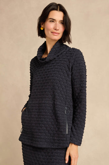 Woman wearing the Origami Pocket Jersey Cowl Neck Top, crafted from textured stretch jersey with long sleeves and zippered pockets, standing against a plain beige background. Made in England.