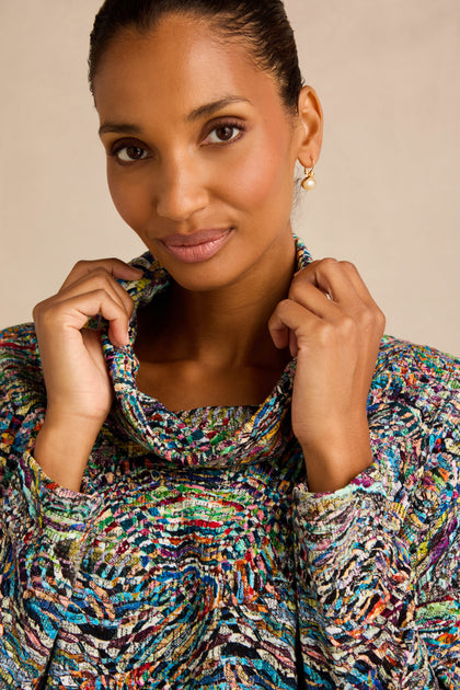 A woman wearing the Patchwork Fields Jersey Top—a colorful, textured top with a boxy silhouette—holds the collar with both hands and looks at the camera with a neutral expression against a plain background.