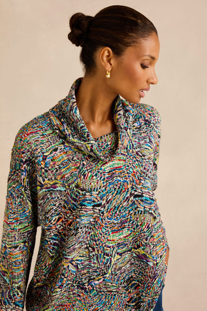 A woman with dark hair in a bun wears the Patchwork Fields Jersey Top—a colorful, patterned patchwork jersey with a cowl neck—paired with gold earrings. She is looking down to her left against a neutral background.