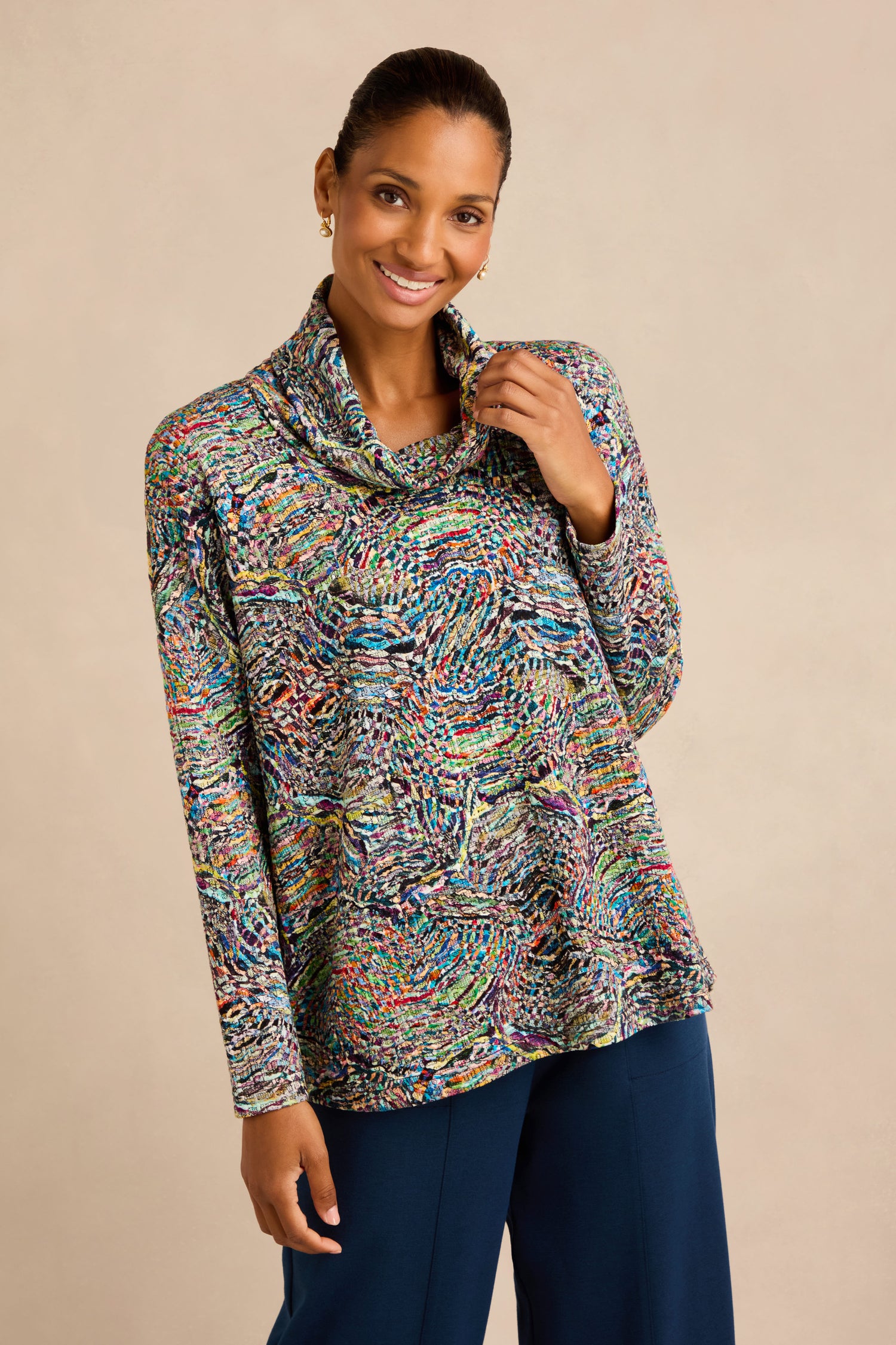 A woman stands against a plain beige background, smiling at the camera in the Patchwork Fields Jersey Top—a multicolored, long-sleeve cowl neck textured jersey with a boxy silhouette—paired with blue pants.