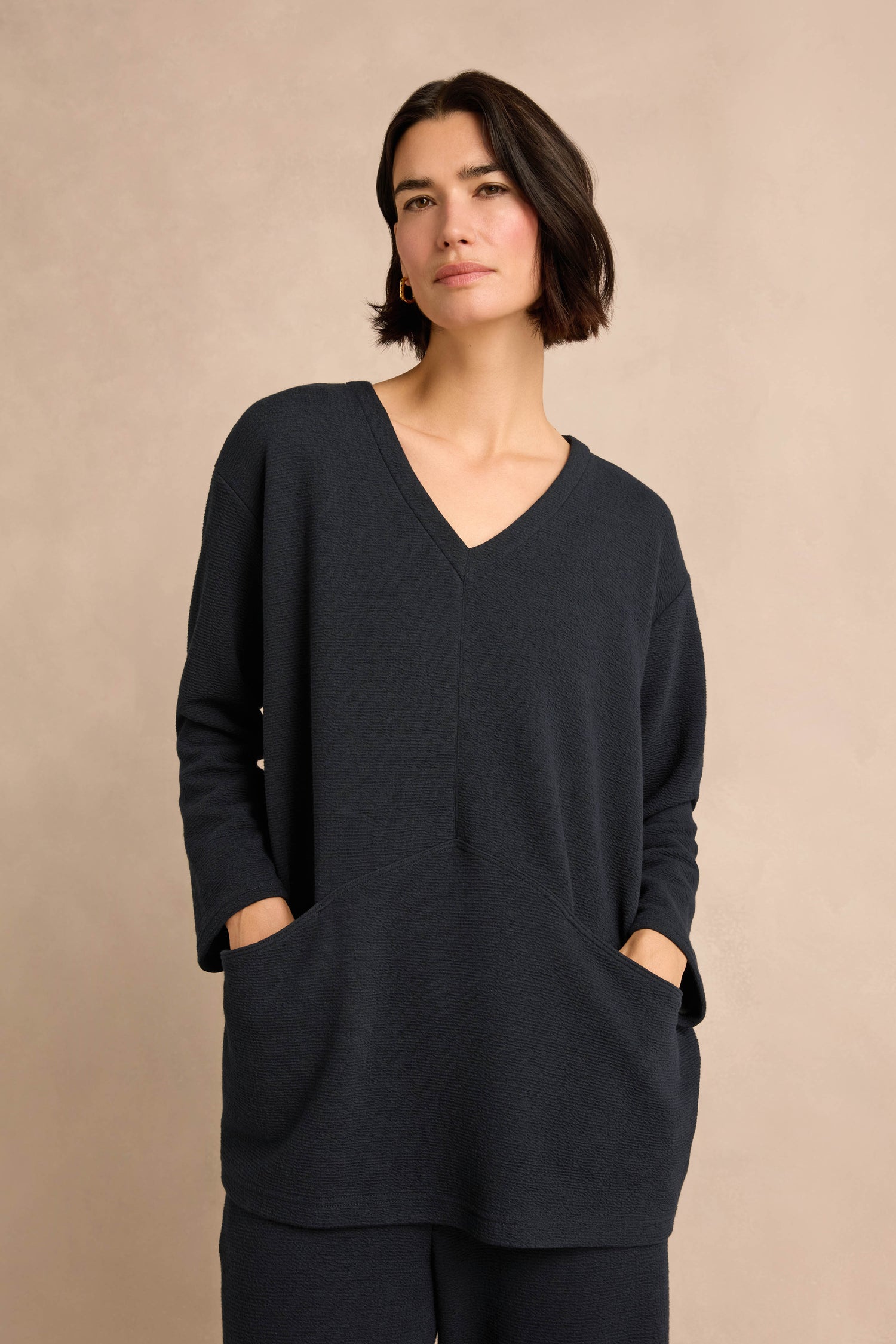 A woman with short dark hair wears the Double Jersey Crinkle Tunic—a loose-fitting, dark tunic sweater with front pockets—paired with matching pants, standing against a neutral background.