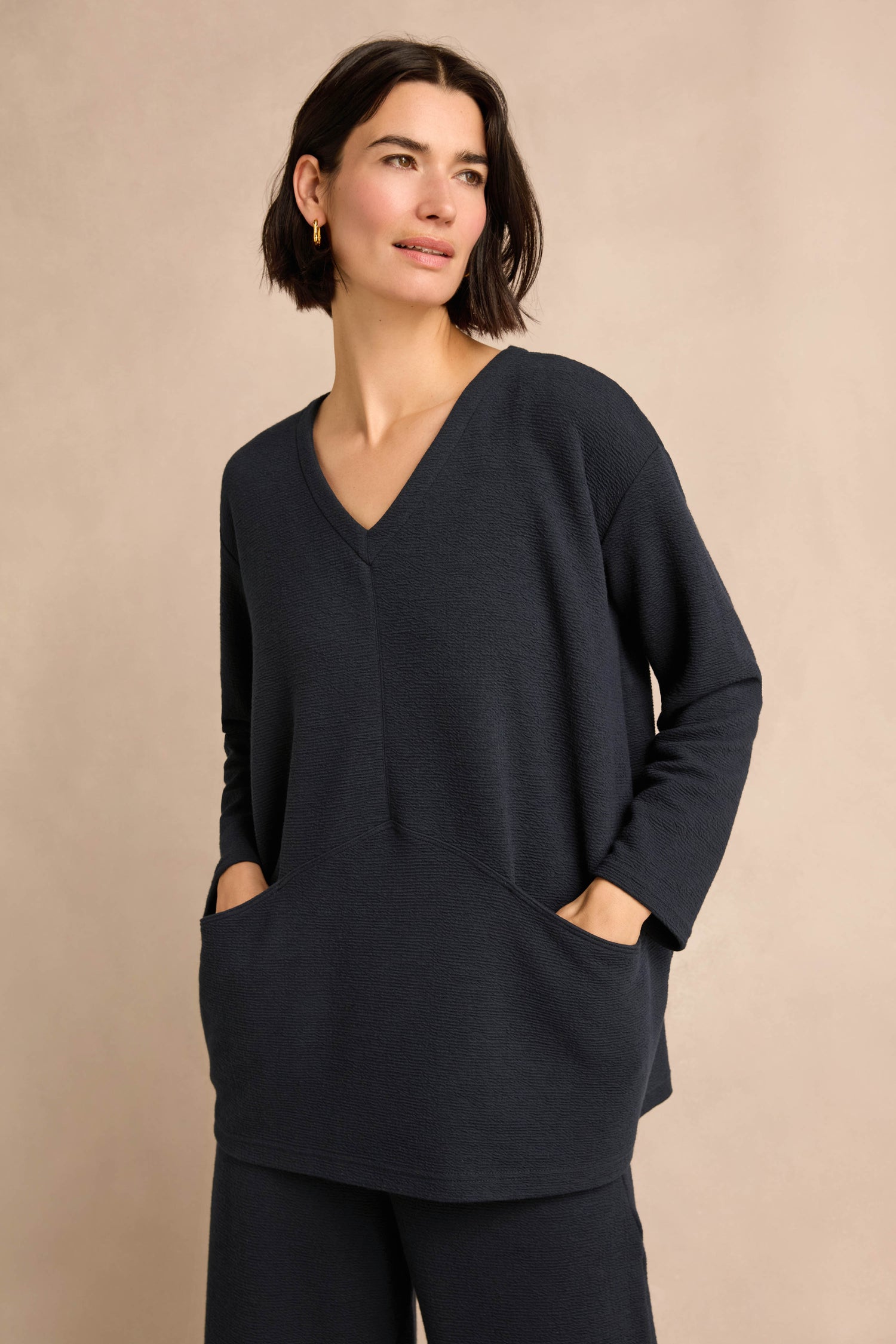 A woman stands against a neutral background wearing the Double Jersey Crinkle Tunic, a loose-fitting, long-sleeved, dark navy style featuring front pockets.
