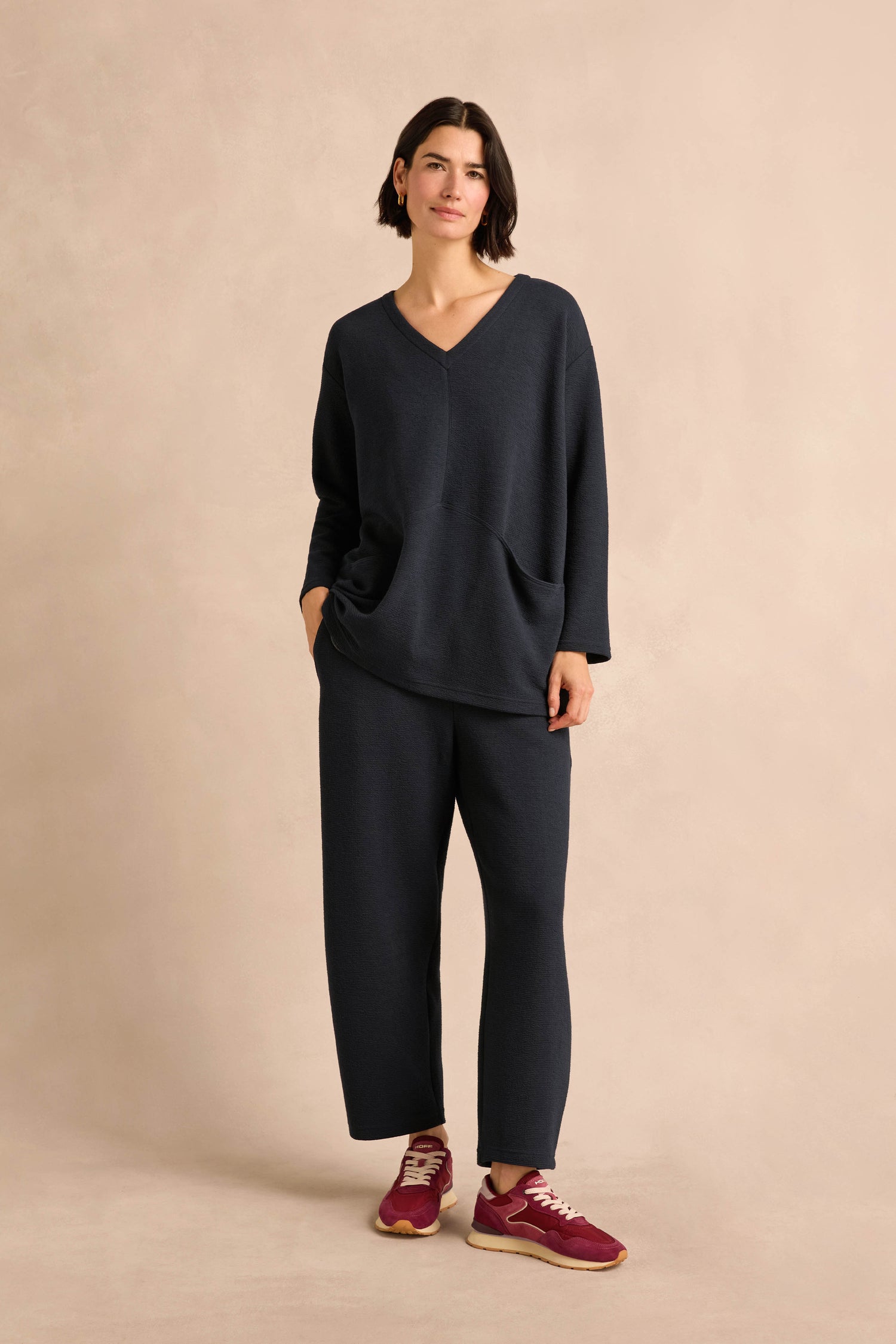 A person stands wearing the Double Jersey Crinkle Tunic in loose-fitting navy, paired with matching pants and maroon sneakers against a neutral beige background. Made in England, this stylish ensemble effortlessly combines comfort with modern elegance.