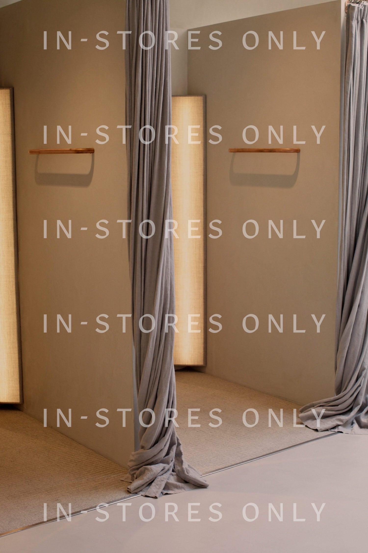 A store changing area with beige walls, grey curtains tied back, and wooden hooks on the walls. Text overlaid reads "IN-STORES ONLY" in white letters. Discover exclusive finds like Bromelia Pendant Earrings, available only here.