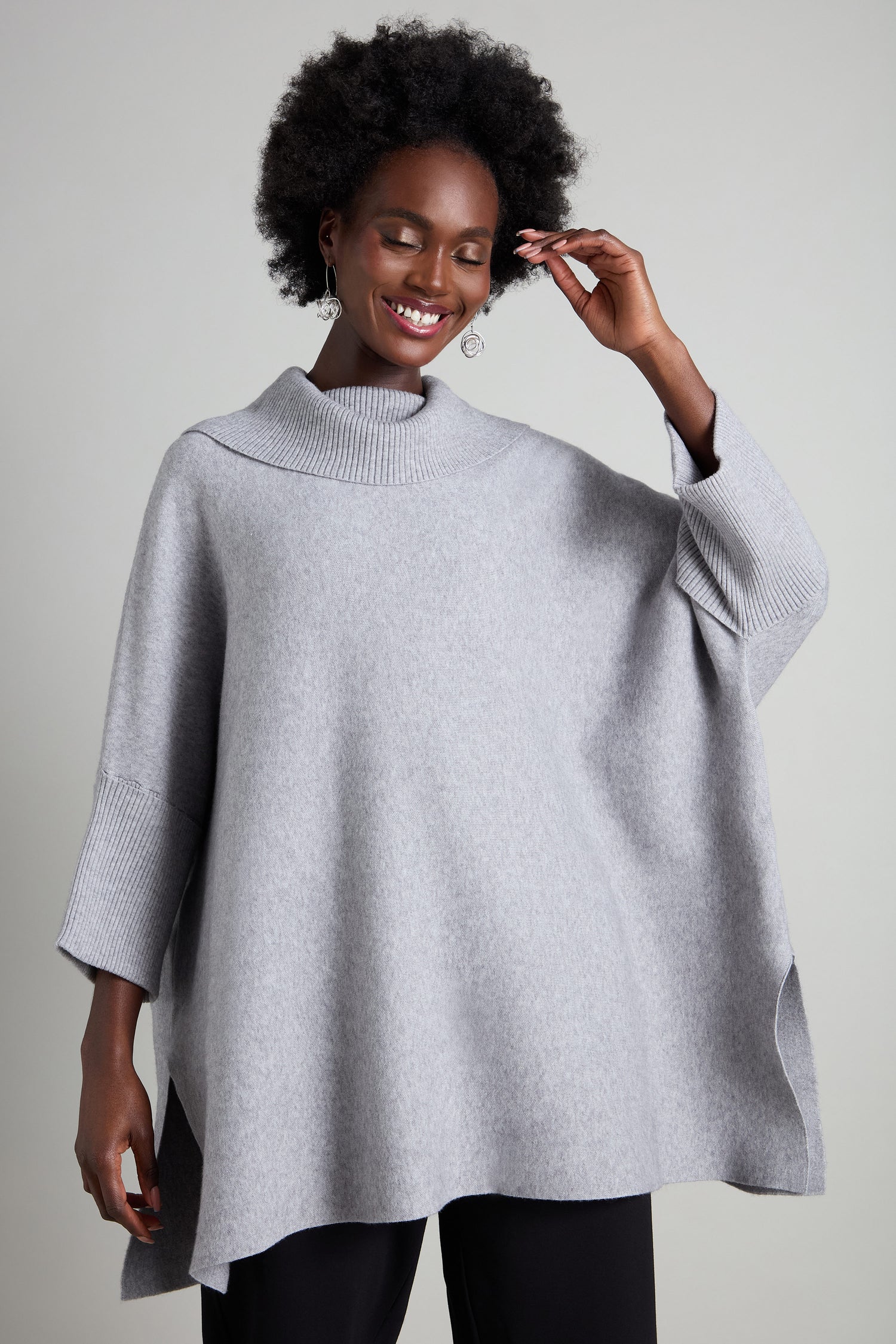 Wearing an oversized sweater called the Roll Neck Knit in gray, a person smiles warmly, posing with one hand gently touching their hair.