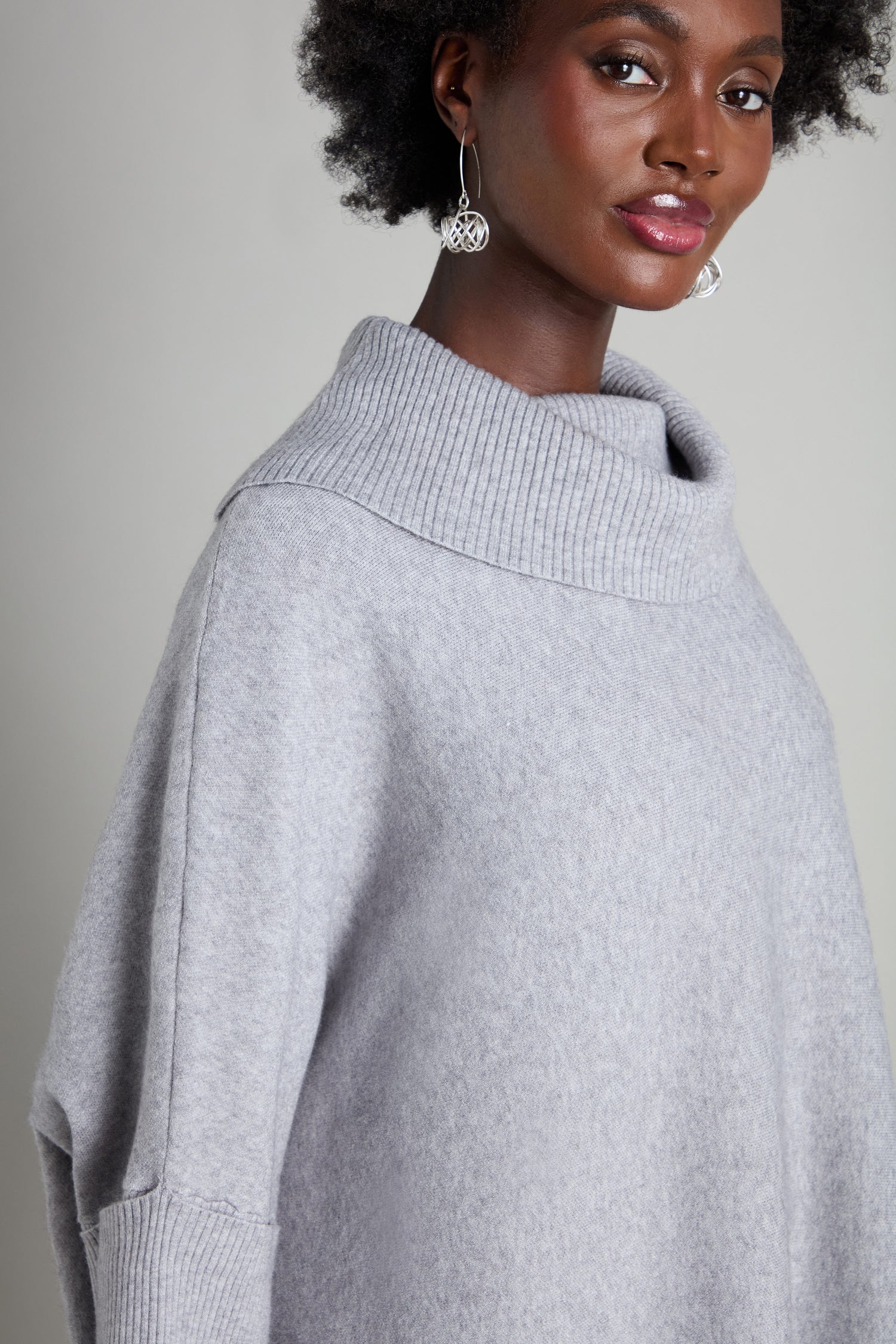 A person dressed in the "Roll Neck Knit" sweater, featuring a gray hue, and adorned with large silver earrings stands against a plain background, showcasing the art of stylish layering.