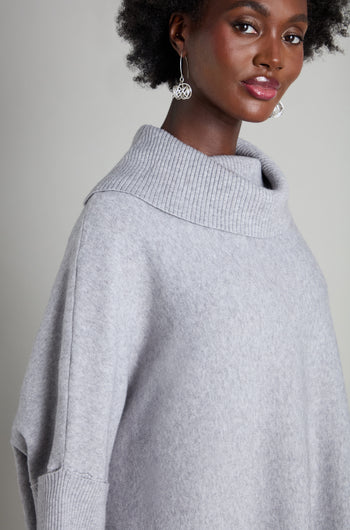 A person dressed in the "Roll Neck Knit" sweater, featuring a gray hue, and adorned with large silver earrings stands against a plain background, showcasing the art of stylish layering.