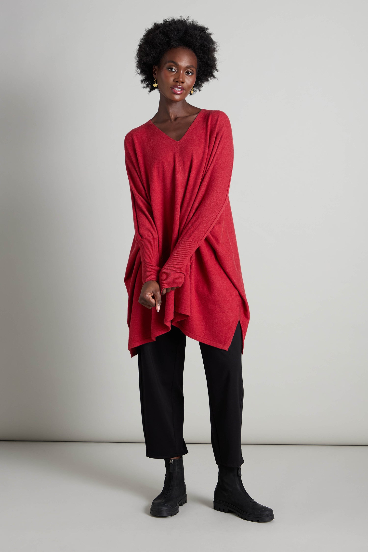 A person wearing a red V Neck Knit, perfect for layering in cooler weather, pairs it with black pants and boots as they stand against a plain backdrop.