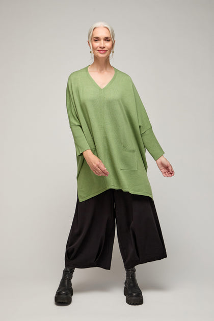A gray-haired woman models the V Neck Knit—a lightweight green top with a pocket—paired with wide black pants and boots against a plain backdrop, embodying minimalist style and versatile, year-round wear.